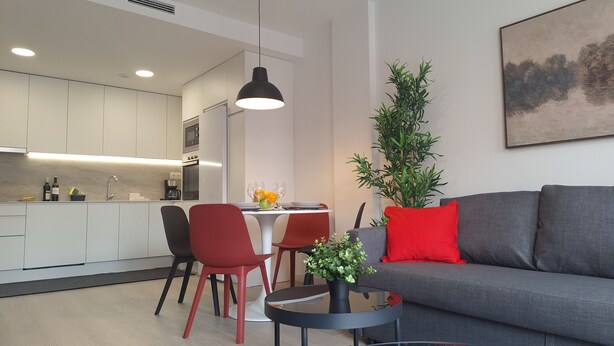 Uma sala de estar moderna com um sofá cinzento, cadeiras vermelhas, uma mesa de jantar com fruta e uma cozinha ao fundo.