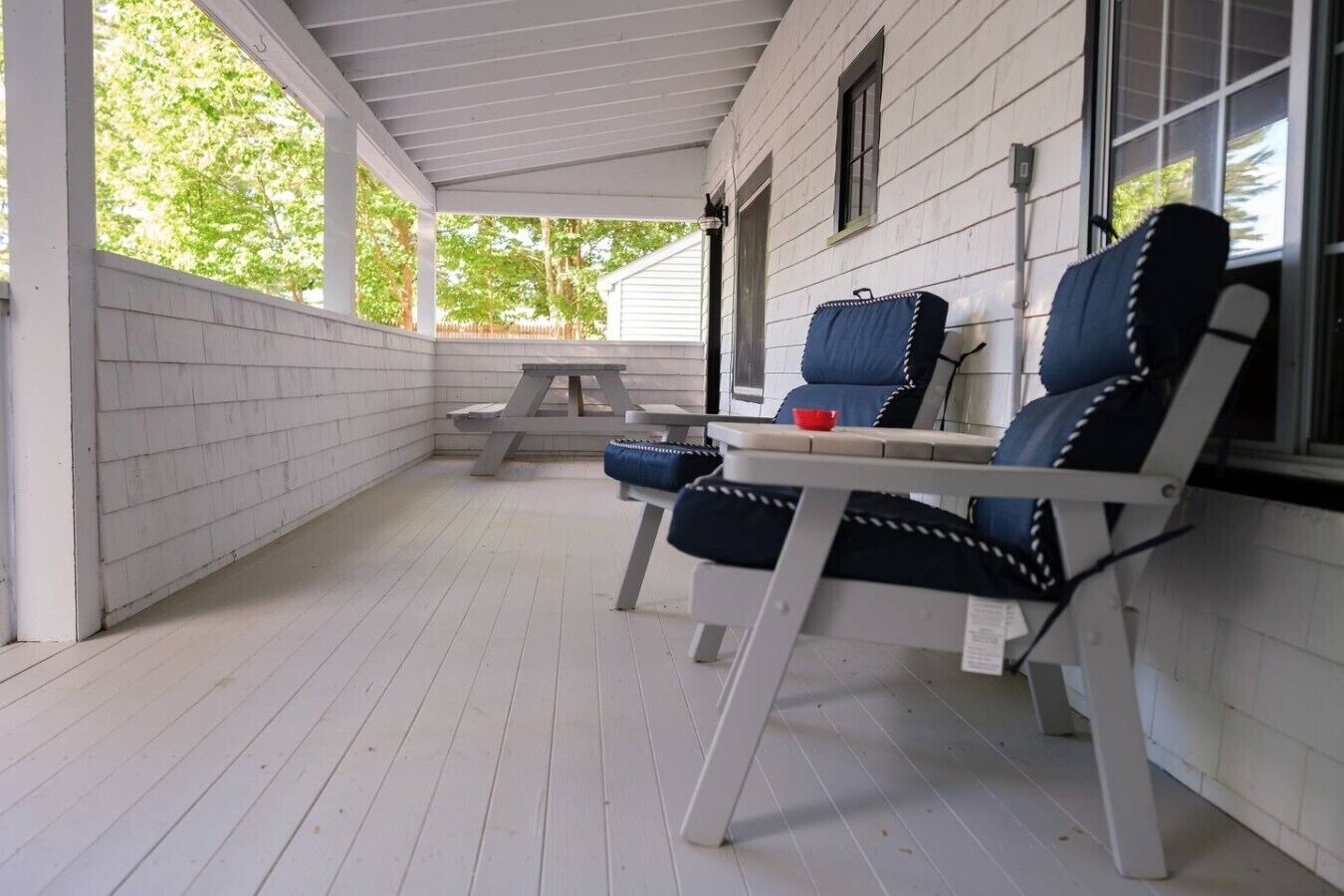 Une véranda couverte aux murs en bois peints en blanc, une table de pique-nique et deux chaises bleues et blanches.