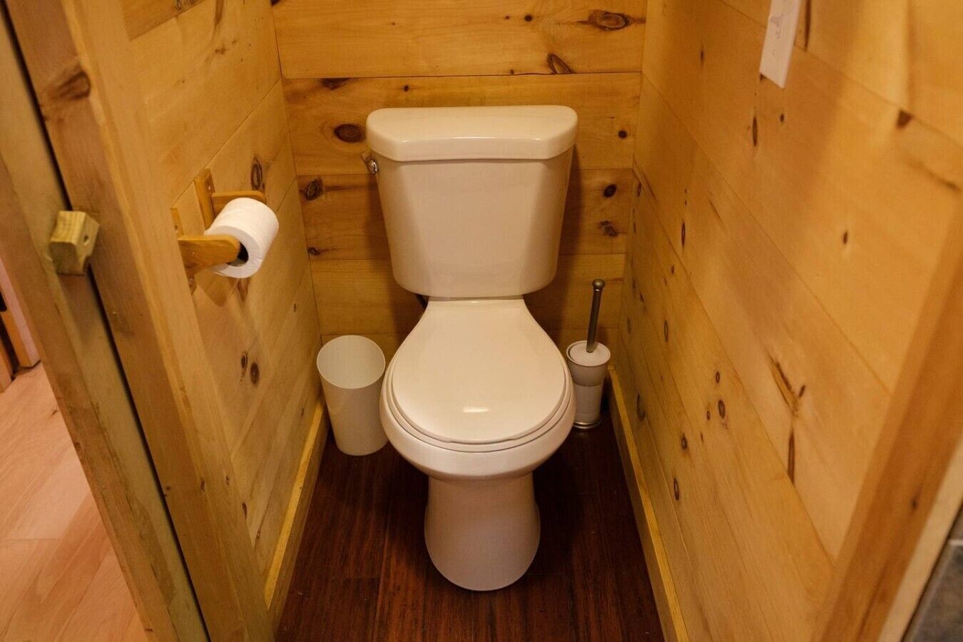 Une salle de bain aux murs lambrissés, équipée de toilettes, d’un porte-rouleau de papier toilette et d’une poubelle.