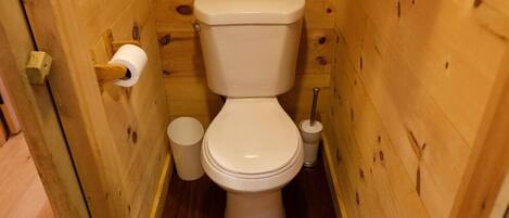 Une salle de bain aux murs lambrissés, équipée de toilettes, d’un porte-rouleau de papier toilette et d’une poubelle.