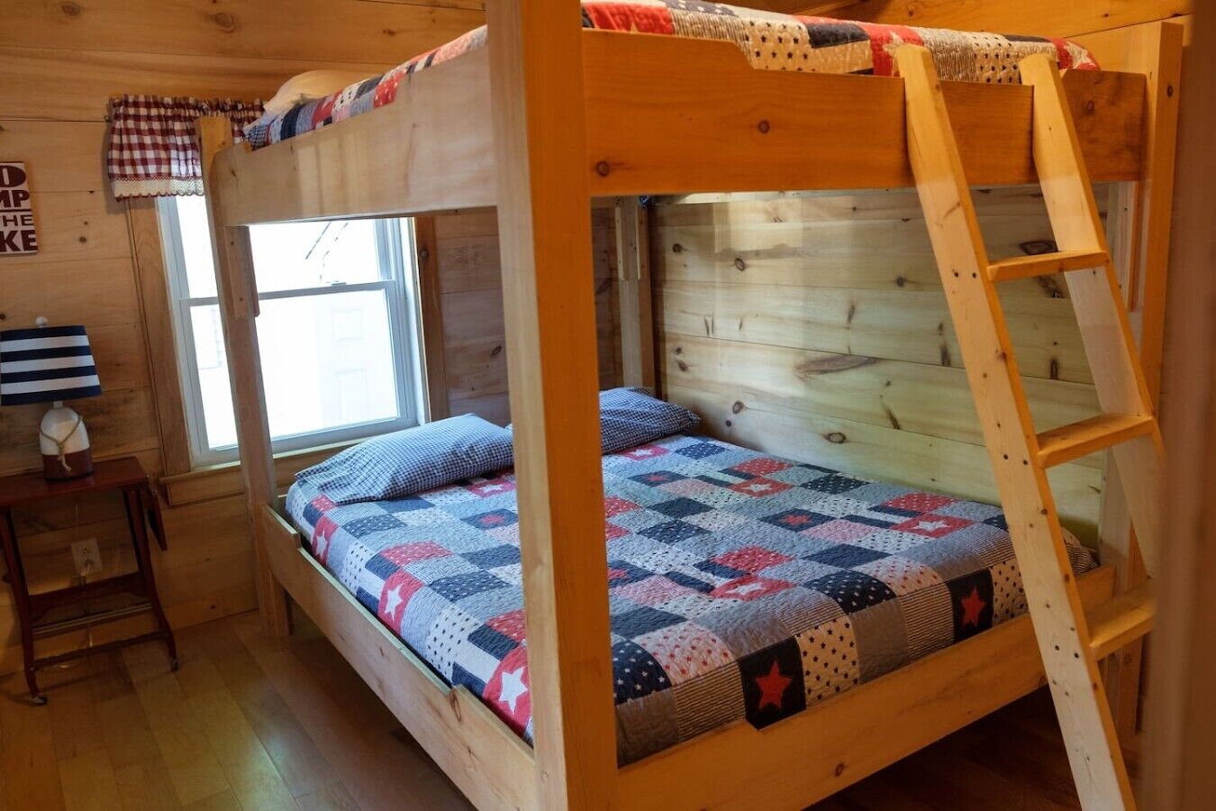 Una litera con escalera, una ventana, una mesita con lámpara y una colcha acolchada.