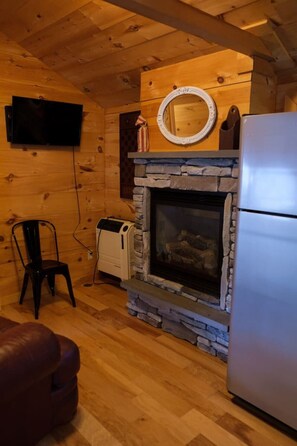 Ein gemütliches Blockhaus-Interieur mit Kamin aus Stein, einem an der Wand befestigten Fernseher, einem Kühlschrank und einem Sessel.