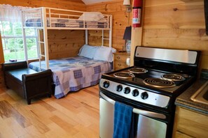 Een houten interieur met een stapelbed, een kitchenette met fornuis, een houten vloer en een raam met uitzicht op groen.