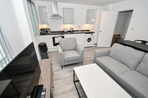A modern kitchen with a sink, stove, and microwave. There is a grey sofa and a small table in the living area. - Contemporary 1 Bedroom Apartment in Leeds (Leeds)