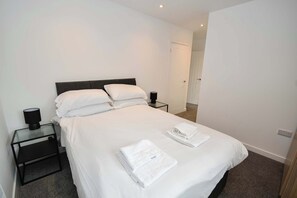 A neatly made bed with white linens, a small bedside table with a lamp, and a folded towel on the bed. - Contemporary 1 Bedroom Apartment in Leeds (Leeds)
