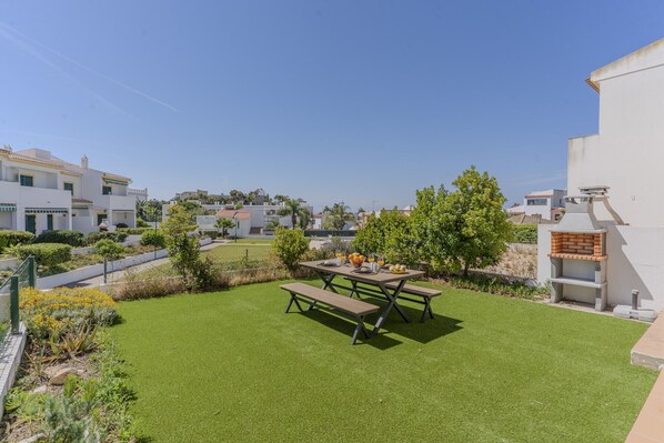 A well-maintained lawn with a picnic table and benches, surrounded by a white building with a brick barbecue area. - #375 Spacious Townhouse with Pool, BBQ, and Parking (Albufeira)