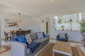 A modern living room with a sofa, coffee table, dining area, and a large window with plants. - #375 Spacious Townhouse with Pool, BBQ, and Parking (Albufeira)