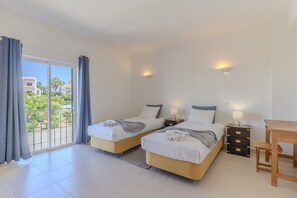 A hotel room with two beds, a wooden table, a nightstand with drawers, and a balcony with curtains. - #375 Spacious Townhouse with Pool, BBQ, and Parking (Albufeira)