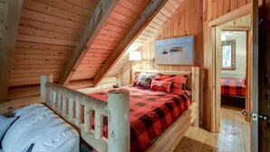 Un dormitorio de cabaña de madera con techo inclinado, una cama con colcha a cuadros rojo y negro, y una ventana con vista a un bote.