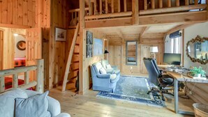 Interior de una cabaña de madera con habitación de altillo, sala de estar con sofá y sillón, escritorio con computadora y mesa de comedor.