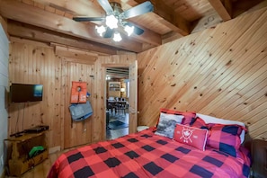 A wooden room with a bed, a ceiling fan, a TV, and a door leading to a dining area. - Lake Muskoka Escape - McVittie Island|Wifi|Firepit (Bracebridge)