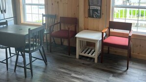 A room with a wooden interior, a television mounted on the wall, a dining table with chairs, and a window with a view of greenery. - Lake Trout Cottage on the Stunning West Shore of Sebago Lake (Sebago)