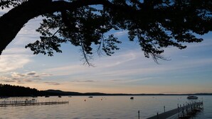 A calm lake with a dock, boats, and a distant shoreline. - Lake Trout Cottage on the Stunning West Shore of Sebago Lake (Sebago)