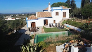 Un edificio blanco con techo de tejas, piscina, y jardín con sillas y mesas.