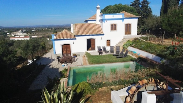 A white building with a tiled roof, a swimming pool, and a garden with chairs and tables.
