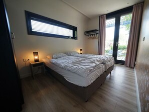 Ein Schlafzimmer mit einem Bett, Nachttischlampen, einem Fenster mit Jalousien und Blick durch eine Glastür.