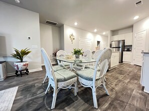 A dining area with a glass table surrounded by white wicker chairs, a potted plant, and a modern kitchen in the background. - Spacious 3BR Family Retreat with Pool Access Near Myrtle Beach (Myrtle Beach)