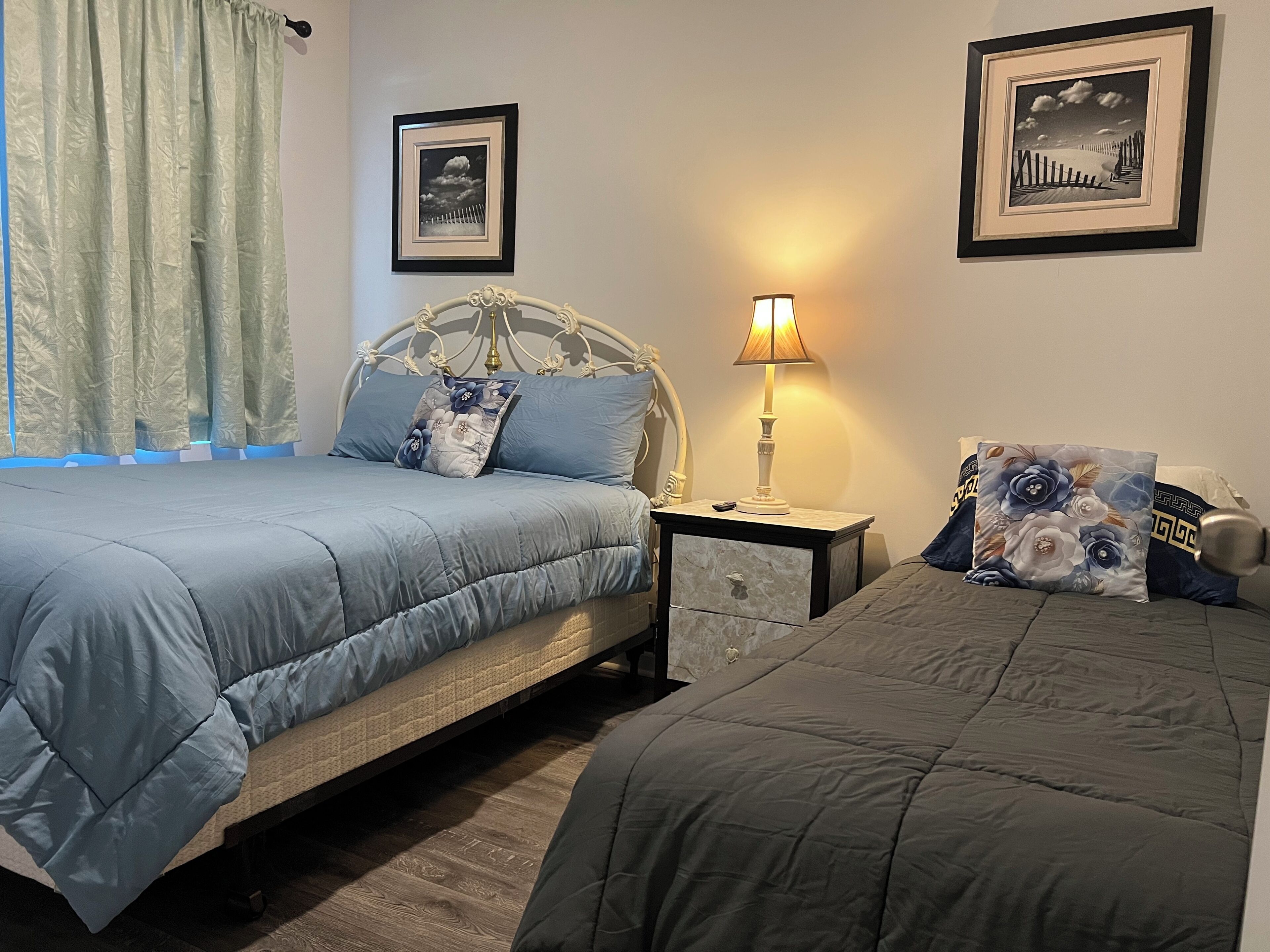 A bedroom with two beds, a nightstand with a lamp, and two framed pictures on the wall.