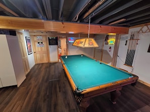 A pool table in a room with wooden flooring, a dartboard, and a neon sign.