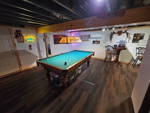 Una mesa de billar en una habitación con suelo de madera, techo con vigas a la vista y varias decoraciones en las paredes, incluyendo un letrero de neón y un cartel.