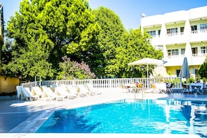 Una piscina con sillas de descanso y sombrillas, rodeada de árboles y un edificio con balcones.