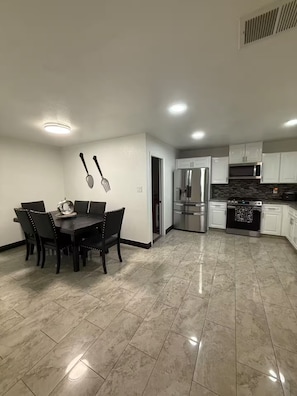 A modern kitchen with a dining area, featuring a stainless steel refrigerator, oven, and microwave, with a tiled floor and white cabinetry. - Anihouse . (Las Vegas)