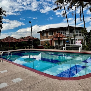 A swimming pool with a tiled deck, a brick building with a red roof, and palm trees.
