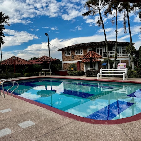 Una piscina con borde de baldosas, un edificio de ladrillo con techo rojo y palmeras.