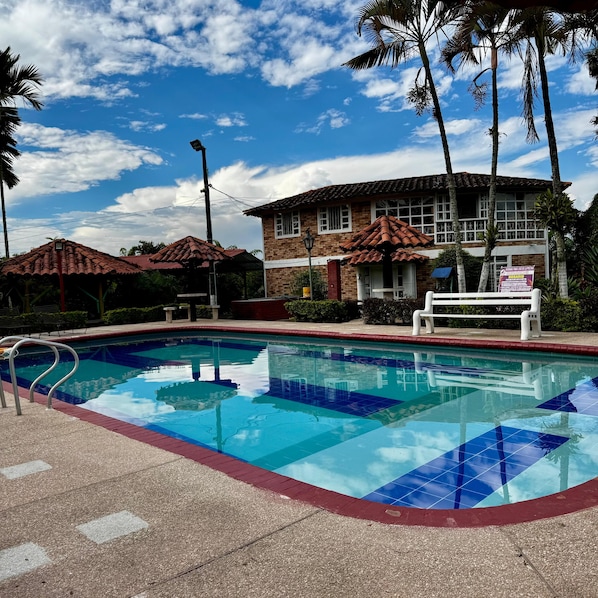 Una piscina con borde de baldosas, un edificio de ladrillo con techo rojo y palmeras.