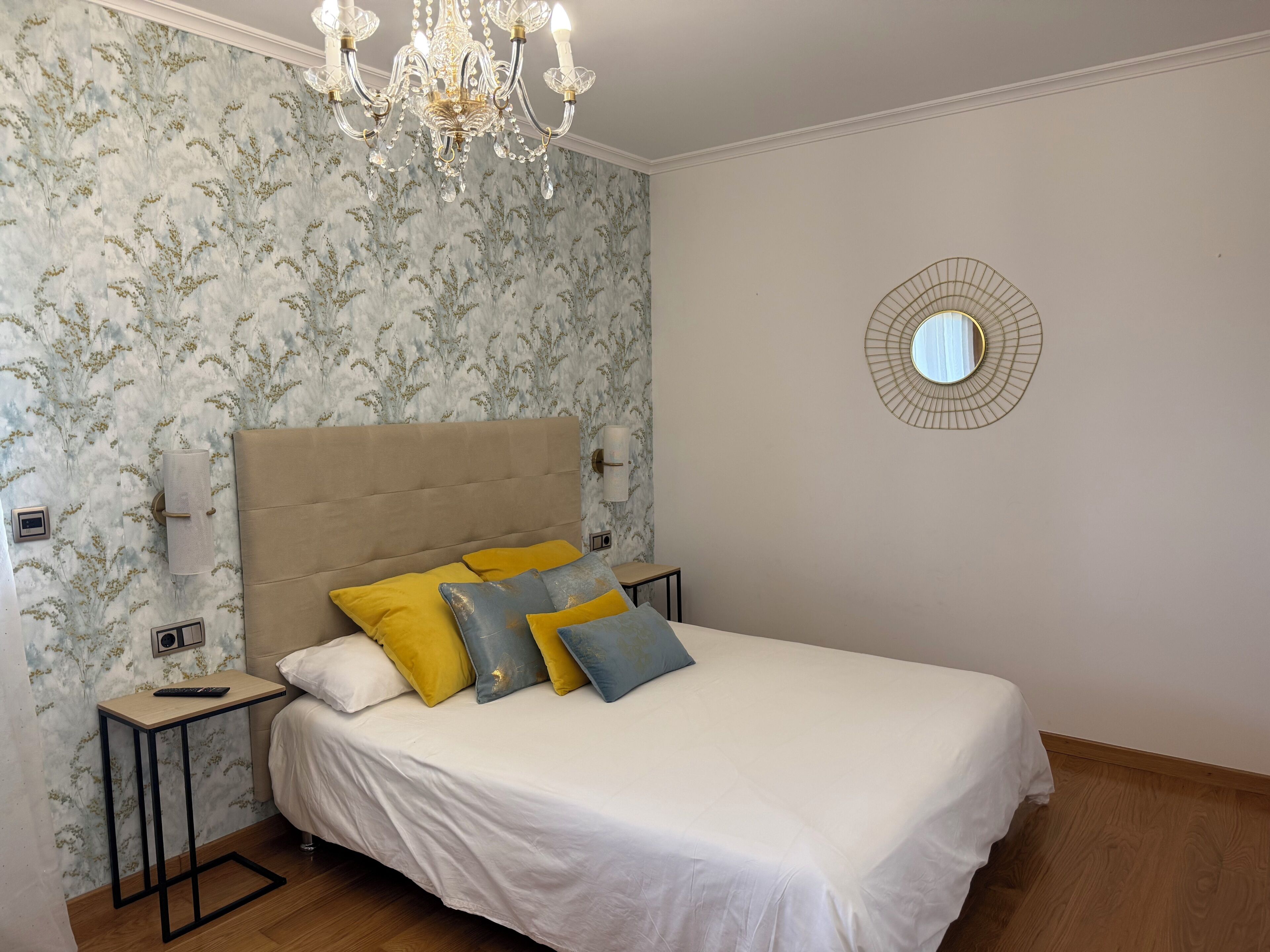 A bedroom with a bed, a chandelier, a wall-mounted mirror, and floral wallpaper.