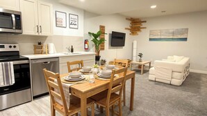 Eine moderne Küche mit weißen Schränken, einer Mikrowelle und einem Geschirrspüler. Es gibt eine Essecke mit einem Holztisch und Stühlen, sowie einen Wohnbereich mit einer Couch und einem Fernseher.