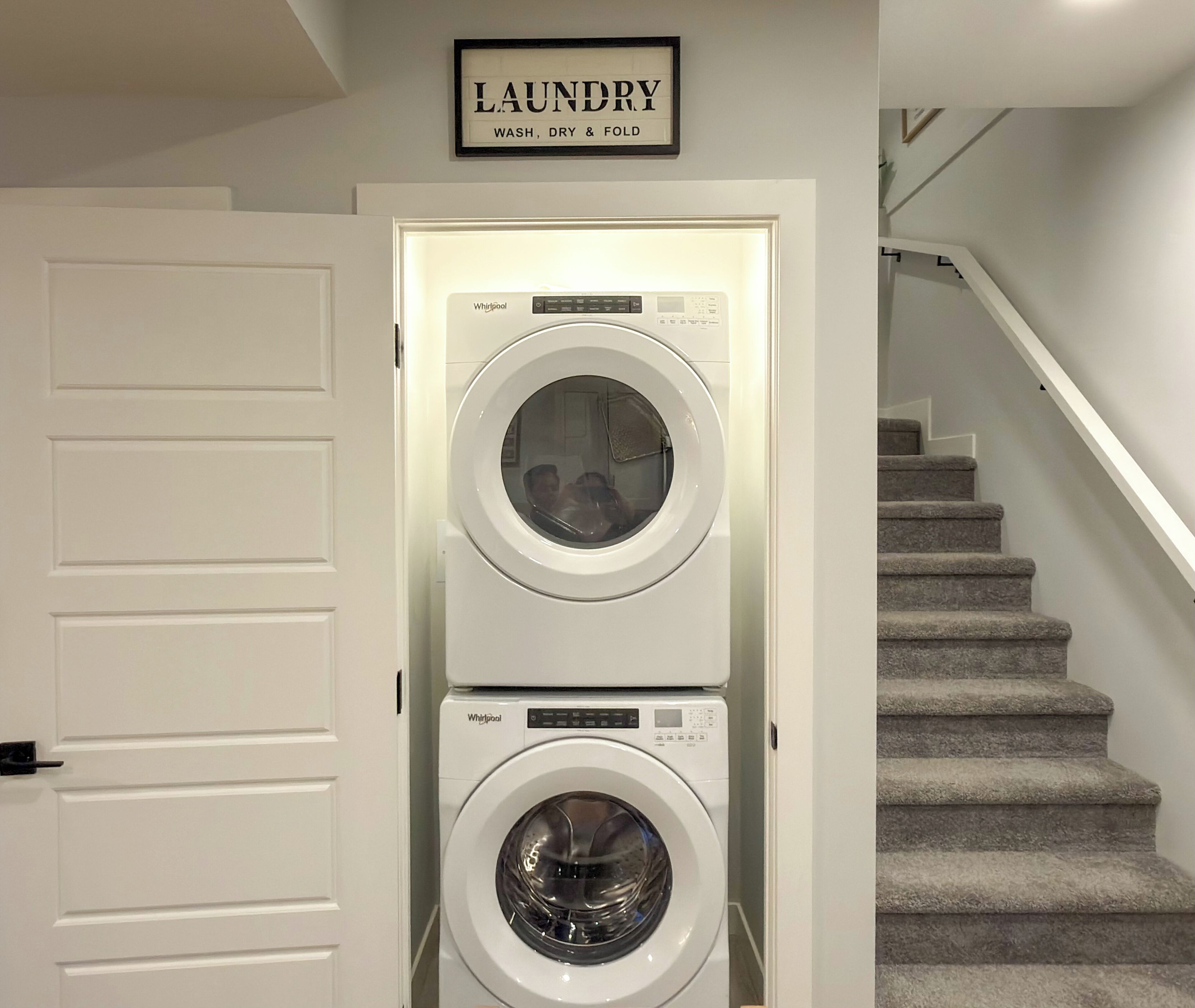 A laundry room with two Whirlpool washers and dryers, a staircase, and a sign above the door that reads 'LAUNDRY WASH, DRY & FOLD.'