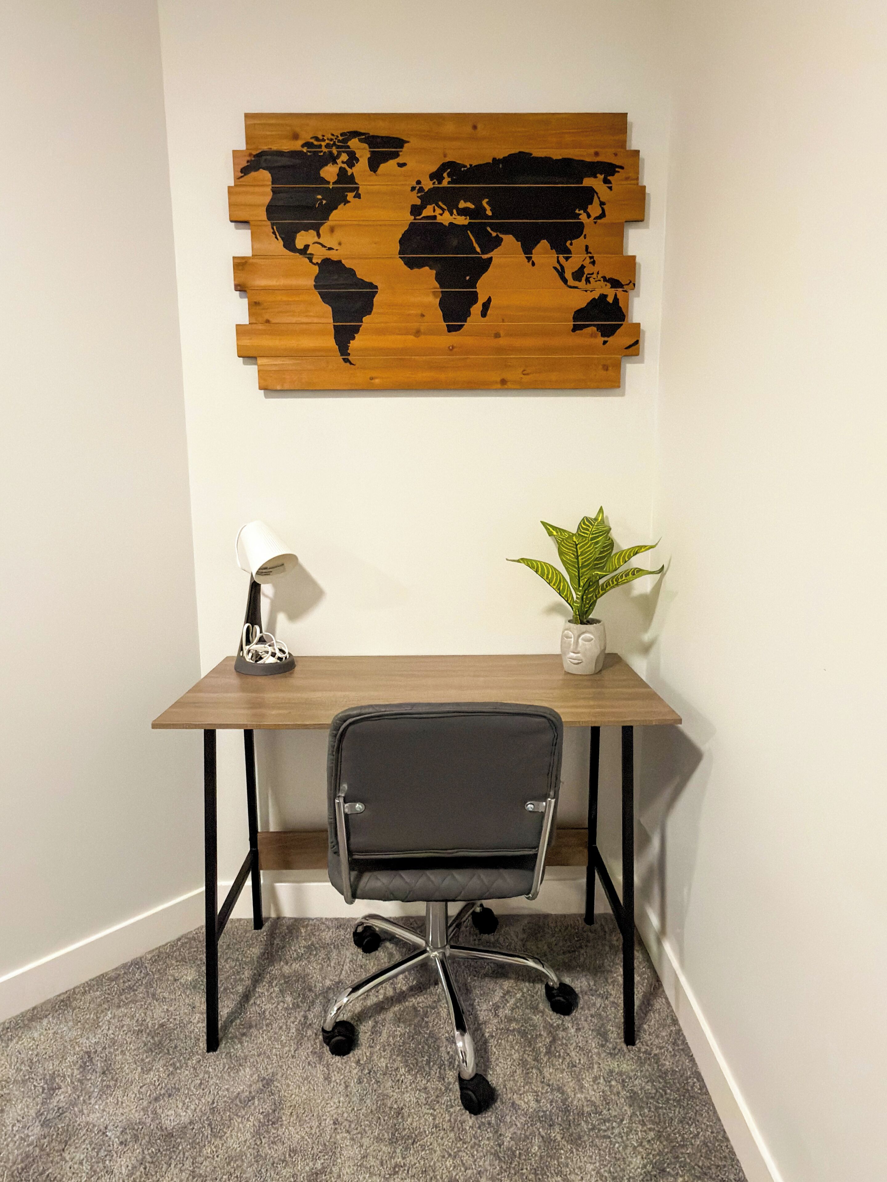 A corner office with a wooden desk, a black office chair, a world map artwork, a white desk lamp, and a potted plant.