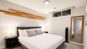 Ein Schlafzimmer mit einem Bett, Nachttischen, einem Spiegel und einem Fenster mit Pflanzen.