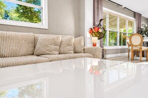 Ein modernes Wohnzimmer mit einem beigefarbenen Sofa, einem Essbereich mit Holztisch und -stühlen sowie einem großen Fenster mit Blick auf Grünflächen.