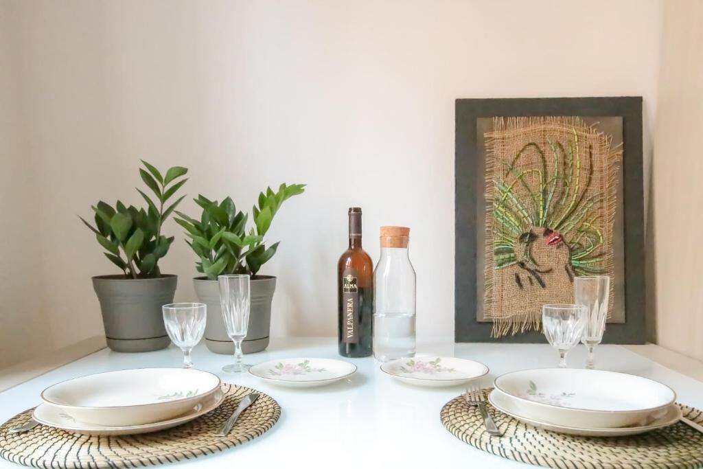 A dining table set with plates, glasses, and bottles, flanked by potted plants and a framed artwork.