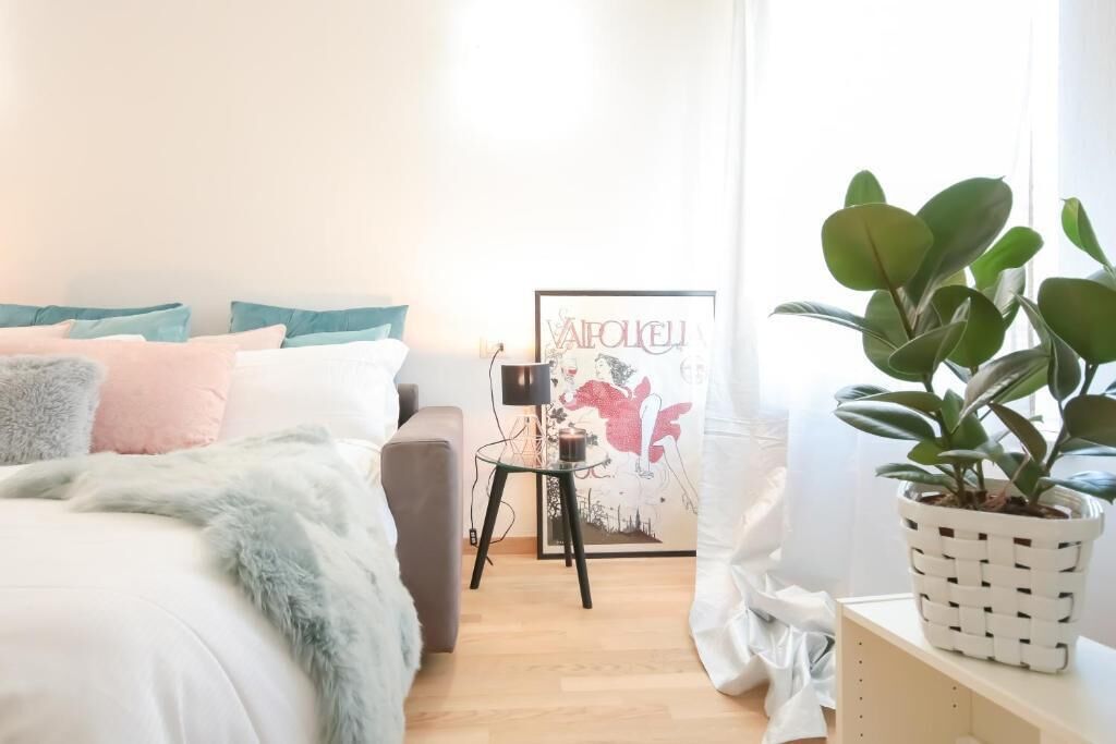 A bedroom with a bed, pillows, a side table, a framed picture, and a potted plant.