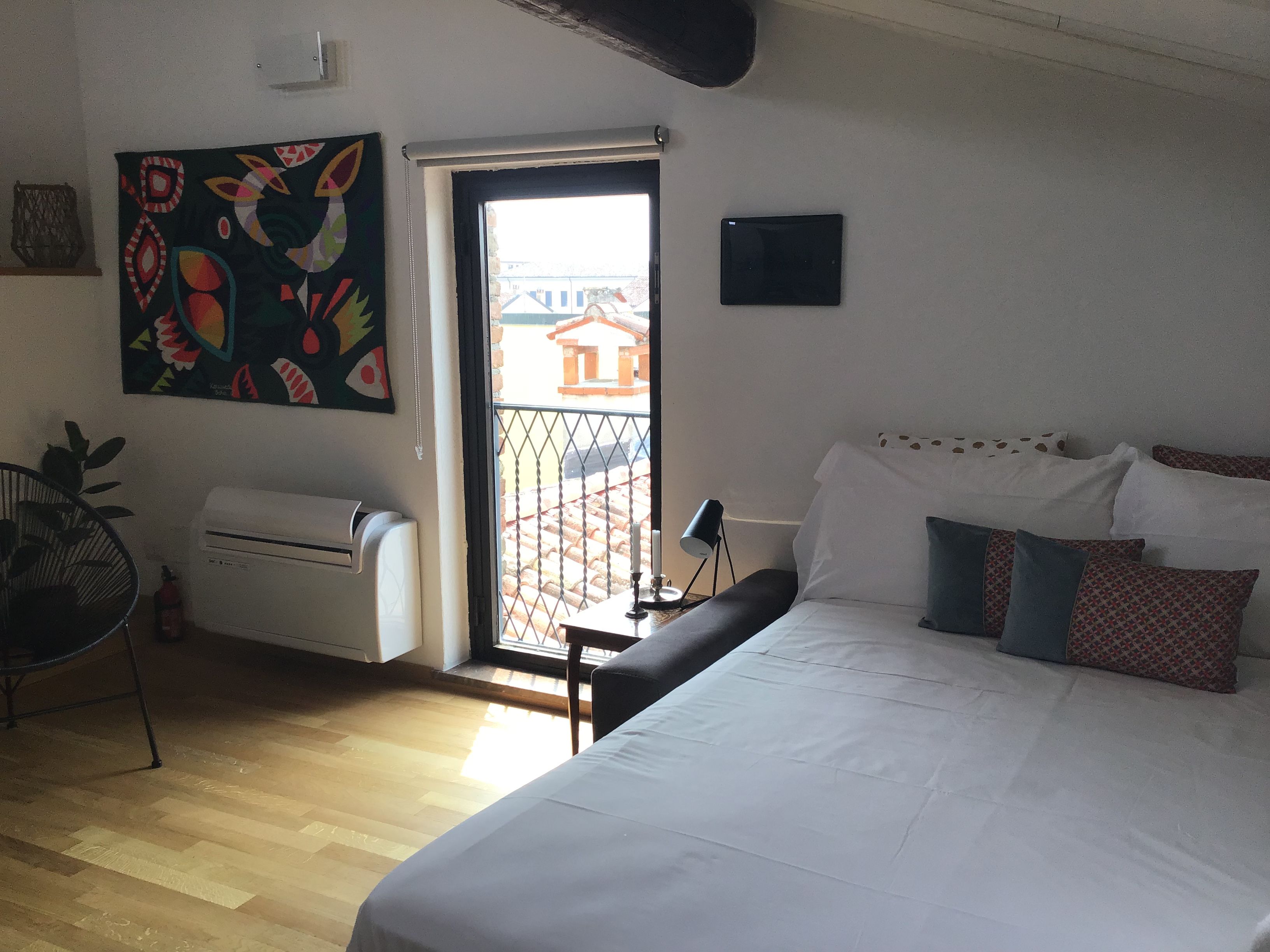 A bedroom with a bed, a window, a door, a radiator, a plant, and a wall-mounted artwork.