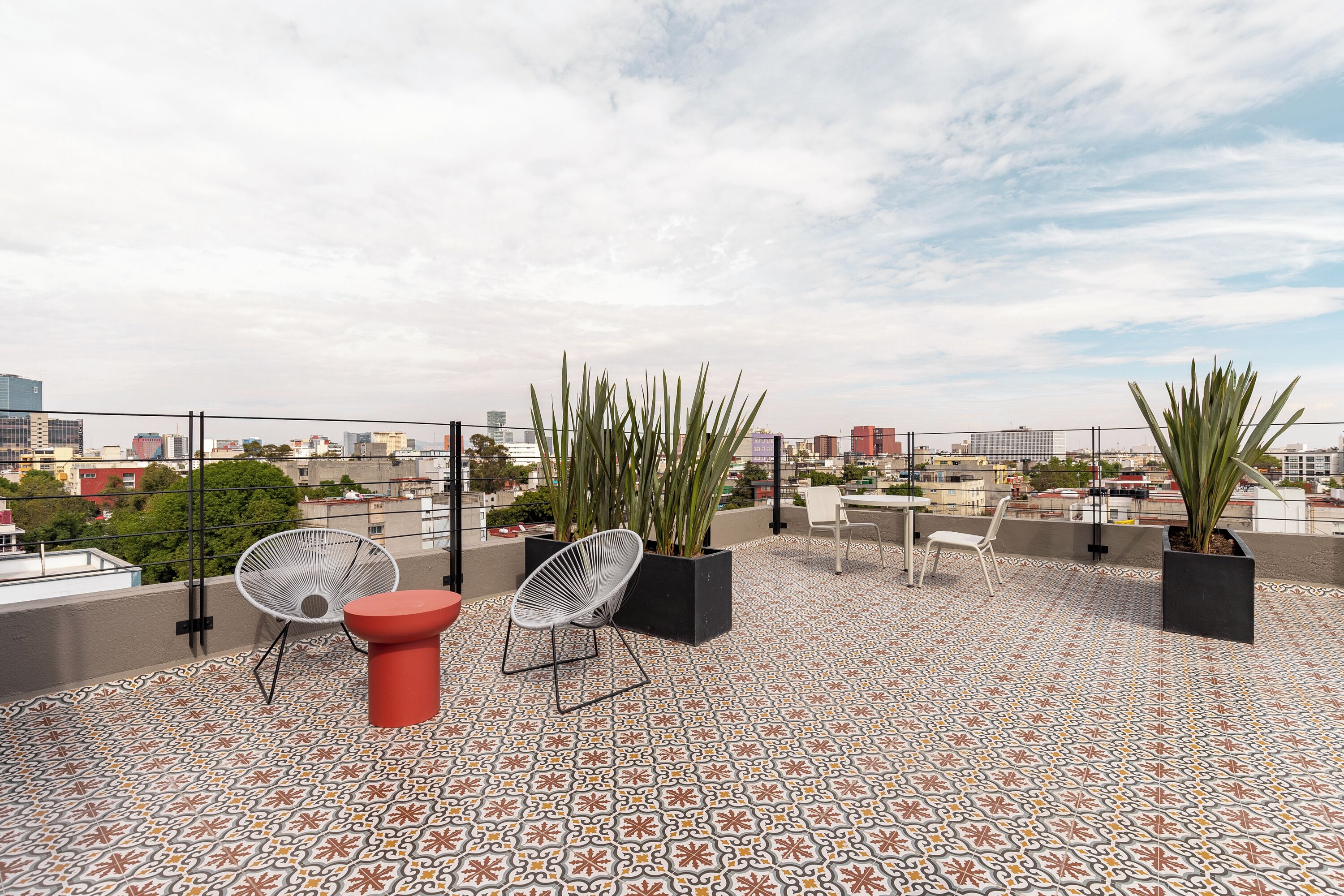 Une terrasse sur le toit avec un sol à motifs, deux chaises blanches, un tabouret rouge et des plantes en pot.
