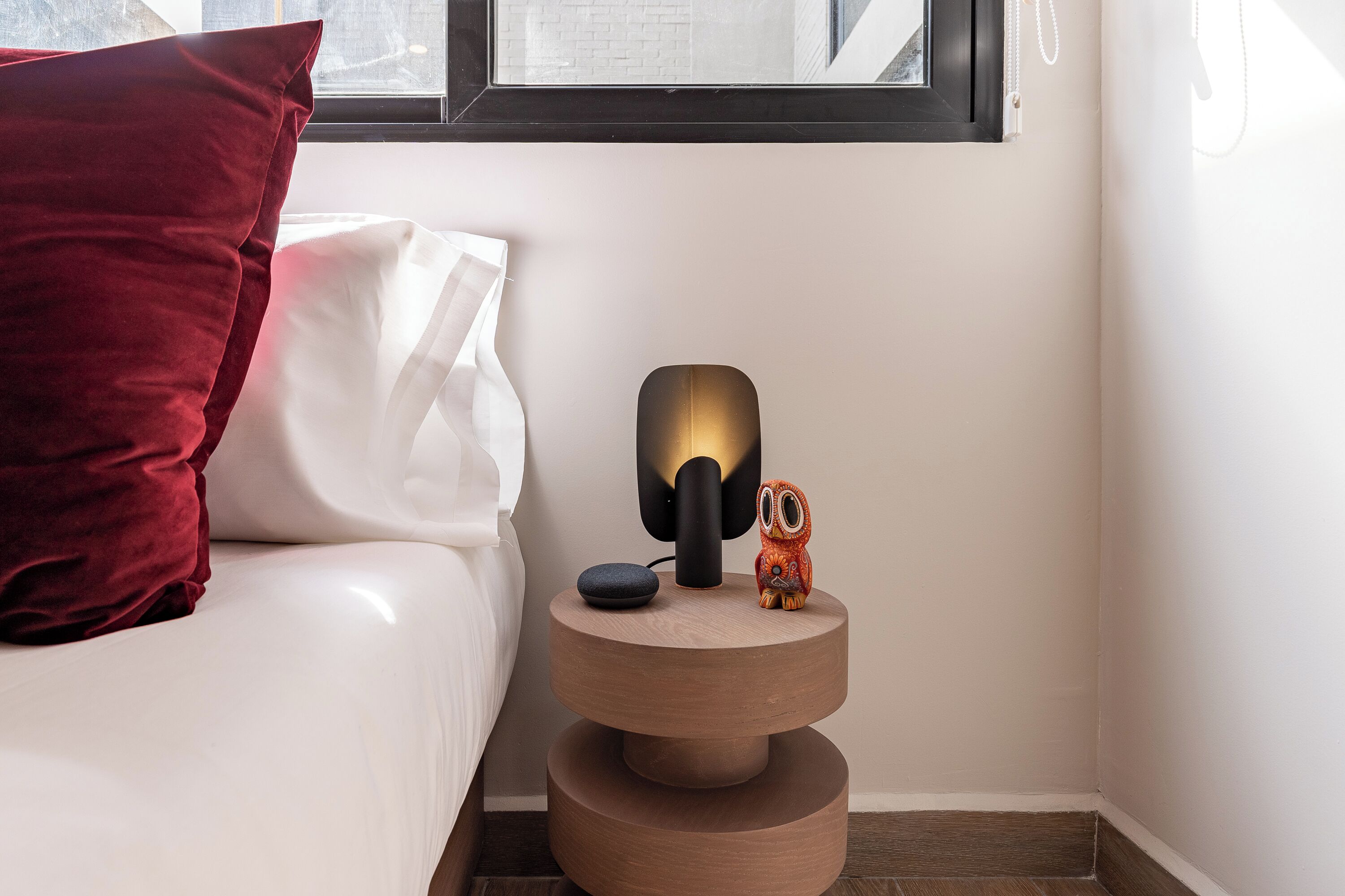 A neatly made bed with a red pillow and a white pillow, a small wooden nightstand with a lamp and a small figurine, and a window above the nightstand.