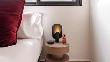 A neatly made bed with a red pillow and a white pillow, a small wooden nightstand with a lamp and a small figurine, and a window above the nightstand.