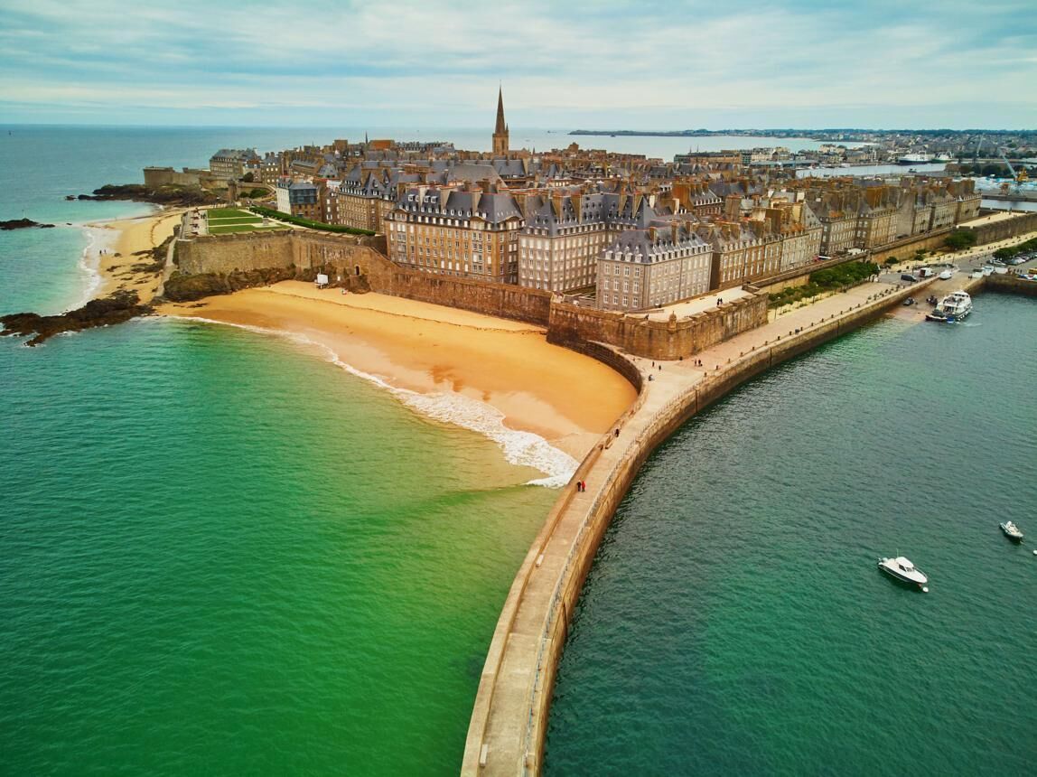 An aerial view of a coastal city with a prominent castle, a sandy beach, and a fortified wall.