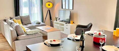 A modern living room with a sofa, dining table set for two, a TV, and a lamp.
