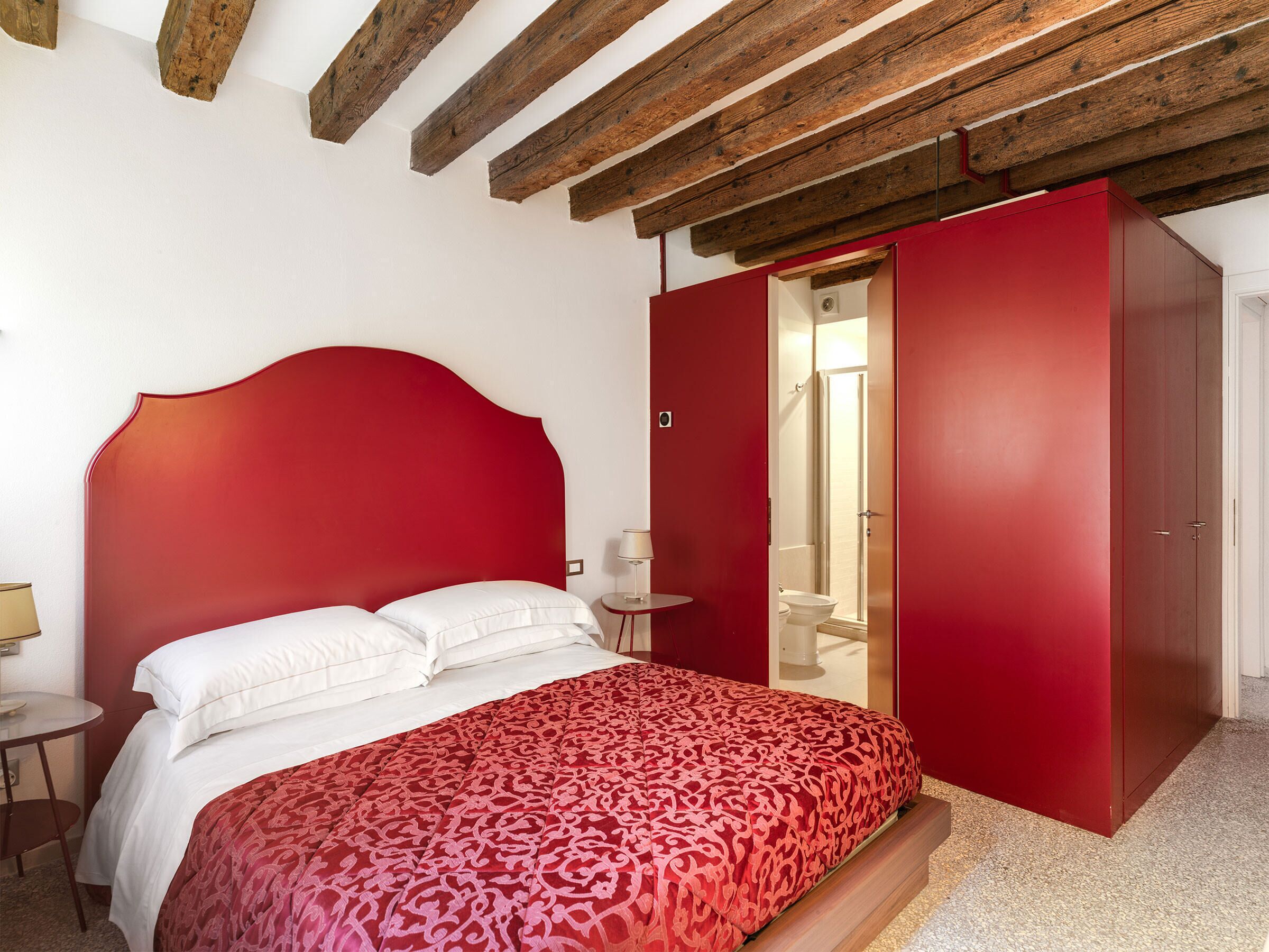 Un dormitorio con cabecera roja, cama con ropa blanca, armario rojo y una mesita de noche pequeña con una lámpara.