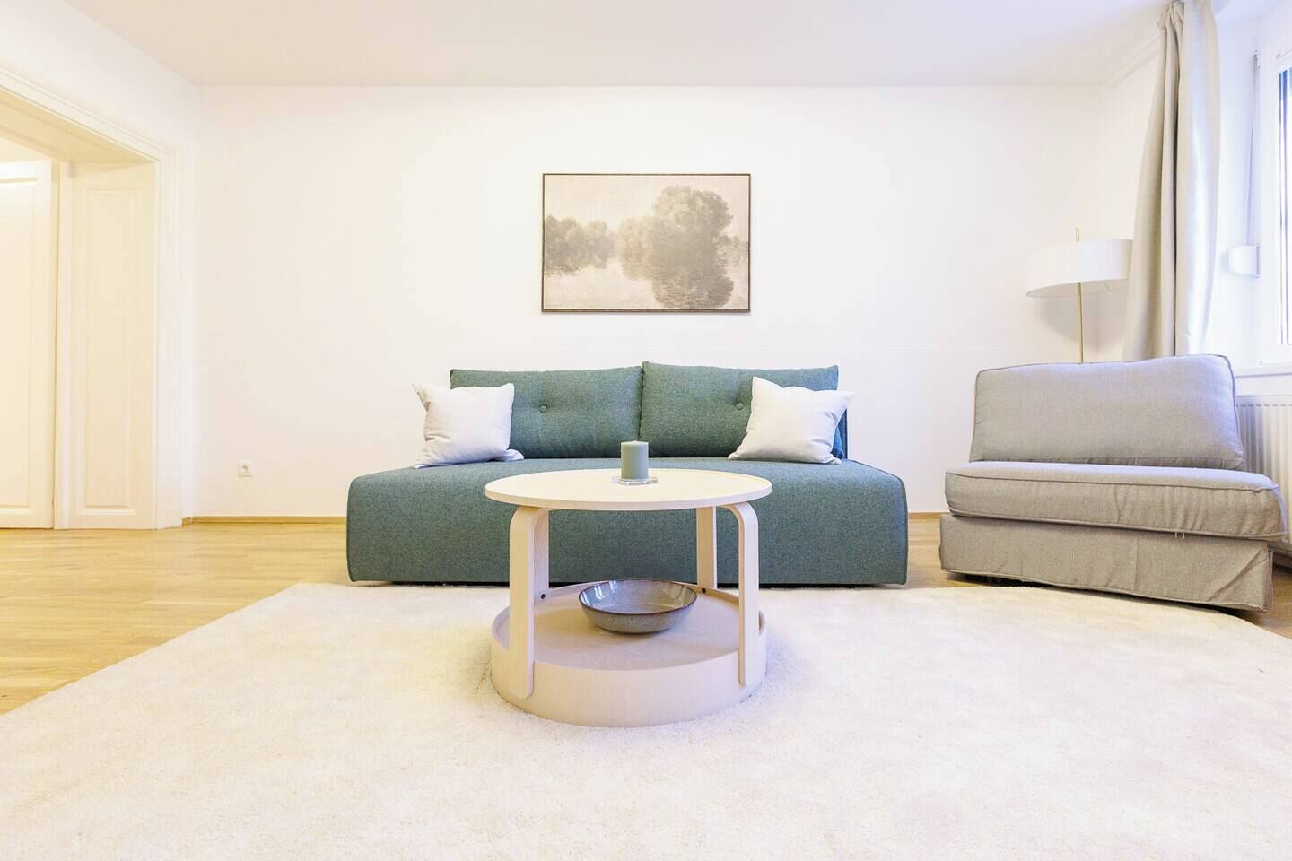 A modern living room with a sofa, a smaller armchair, a round coffee table, and a painting on the wall.