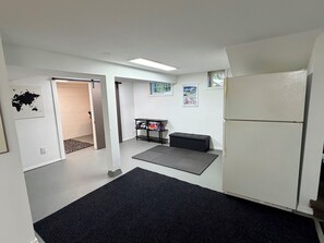 Ein kleines Zimmer mit Kühlschrank, einem kleinen Tisch mit Büchern, einer Matte auf dem Boden und einer Tür zu einem anderen Raum.