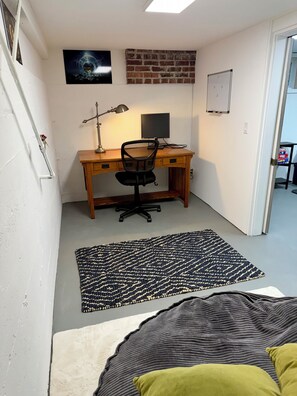 Ein Zimmer mit Schreibtisch aus Holz, einem Computer, einem Stuhl, einem Teppich und einem Bett mit Kissen.
