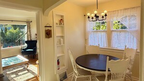 Uma sala de jantar com uma mesa redonda, cadeiras brancas, um candelabro, cortinas de renda e vista para o exterior.