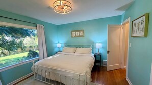 Quarto com cama, abajures, janela com vista para vegetação e porta que dá para outro cômodo.