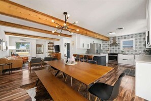 A modern kitchen with wooden beams, a dining area with a long table, and a view of the outdoors.